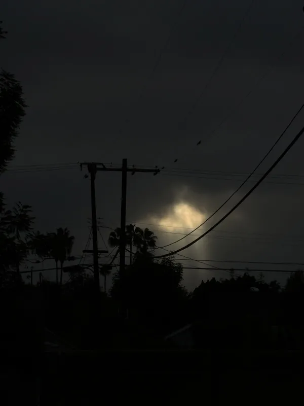Dark sky with clouds, a bright spot with rays of sunlight piercing through. Telephone and electric wires are backlit as well as a few palm trees on the horizon.